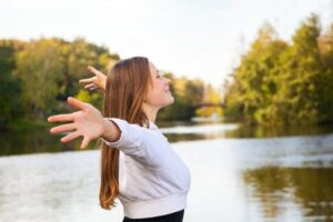 Uśmiechnięta kobieta z szeroko rozłożonymi ramionami nad brzegiem rzeki, biorąca głęboki oddech i uwalniająca napięcie z ciała